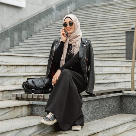 modern stylish muslim woman in hijab, leather jacket and black abaya walking in city street talking on smartphone, wearing sunglasses and bag, fashion style trend